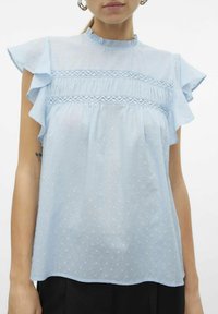 Blusa azzurro chiaro con maniche arricciate, tessuto testurizzato e dettagli in pizzo sul petto. Presenta un delicato motivo a pois.