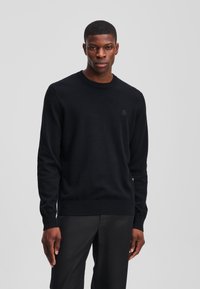 Suéter negro de punto con cuello redondo, puños y dobladillo acanalados, que presenta un sutil logo en el pecho sobre una textura suave. Se lleva con pantalones negros.