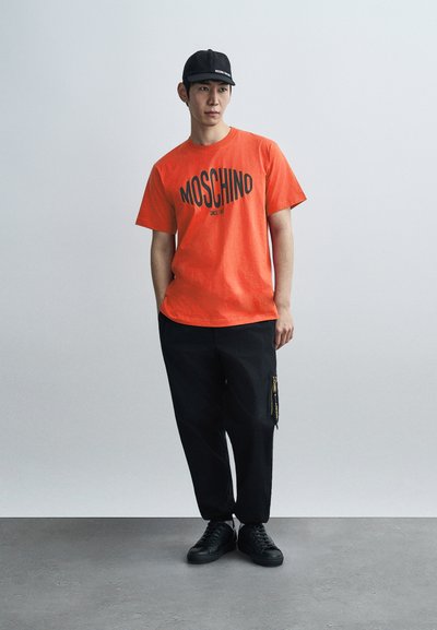 Πορτοκαλί μπλουζάκι Moschino με μαύρο λογότυπο, συνδυασμένο με μαύρο παντελόνι που διαθέτει τσεπάκι με φερμουάρ και μαύρα αθλητικά παπούτσια. Απλός, casual σχεδιασμός.