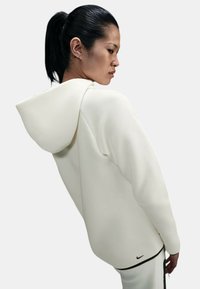 Sudadera Nike en color crema, fabricada con material suave. Presenta una gran capucha, un diseño elegante y un sutil acento del logo negro en el dobladillo inferior.