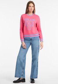 Růžová mikina s modrými detaily a logem "Guess Jeans", doplněná širokými modrými džíny. Hladká textilie a uvolněný design.