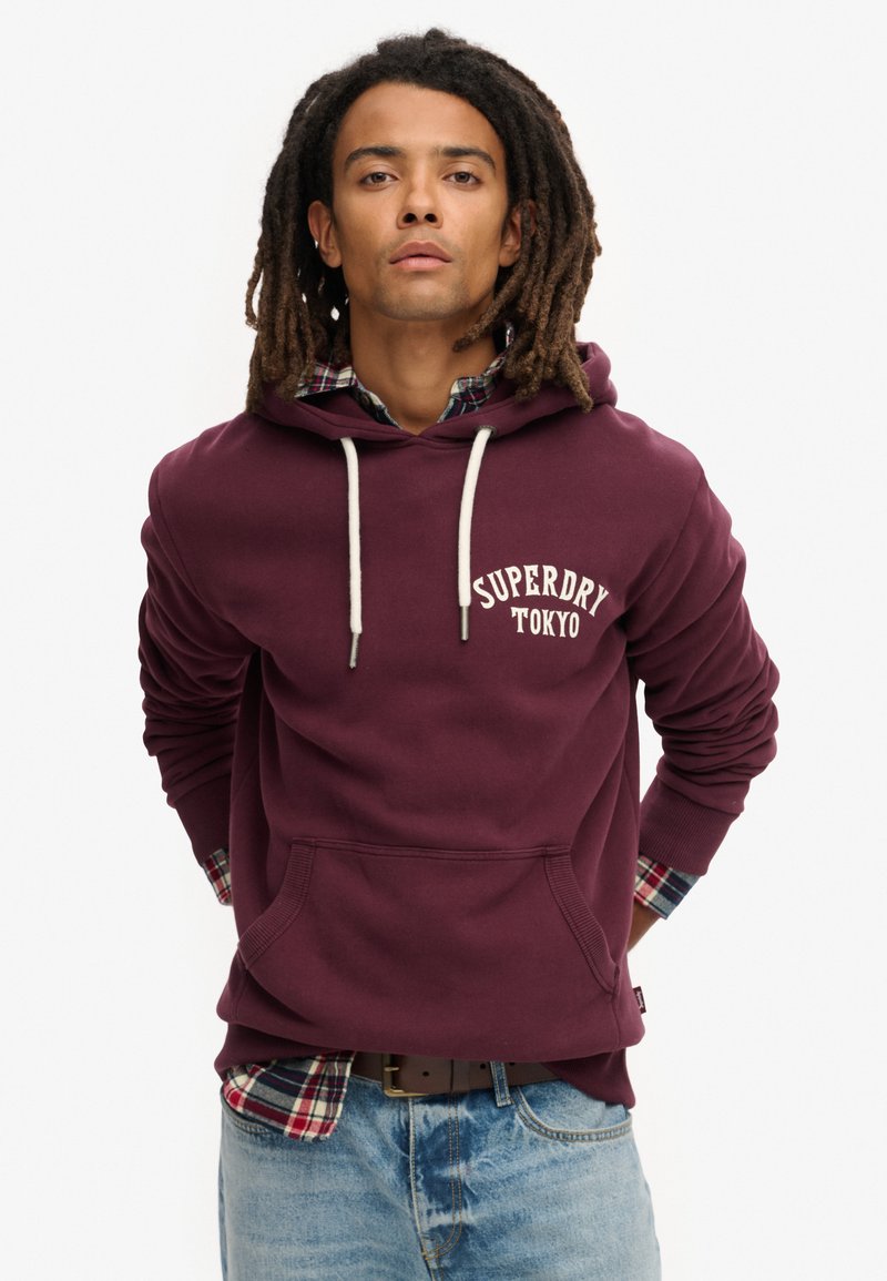 Felpa cappuccio bordeaux con lacci bianchi, tasca frontale e logo "Superdry Tokyo". Stratificata con una camicia a quadri al colletto.
