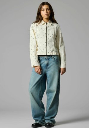 Jeune femme debout, portant une veste blanche zippée avec des points noirs et un jean large bleu clair sur un fond gris.