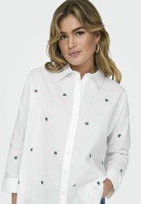 Valge nööpidega särk, millel on rohelise südamekujuline tikand, pikkade varrukatega, millel on keeratud mansetid, ja klassikaline kraedi design.