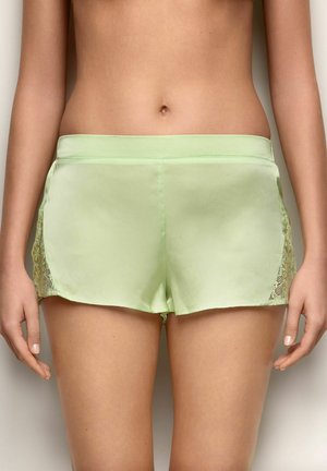Donna che indossa pantaloncini di raso verde chiaro con pannelli di pizzo sui lati, mostrata dalla vita fino alla parte superiore delle cosce, in piedi contro uno sfondo semplice.