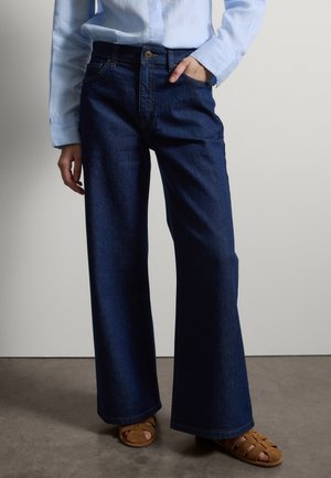 Persoon draagt wijde donkere blauwe jeans, lichtblauw overhemd met knopen en beige sandalen met open tenen, staand tegen een effen lichte muur.