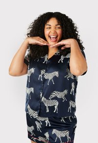 Satängpyjamasuppsättning med marinblå bakgrund och vit zebramönster. Topp med kort ärm och knappfram, samt matchande shorts. Röda accenter.