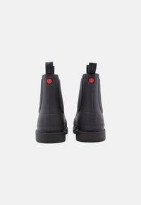 Μαύρες καουτσούκ μπότες αστραγάλου με λεία υφή, με κόκκινη λεπτομέρεια λογότυπου και τράβηγμα για εύκολη τοποθέτηση, τοποθετημένες σε μια ανθεκτική σόλα.
