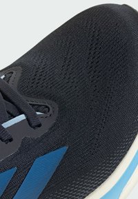 Zapato deportivo negro con parte superior de punto texturizado, acentos de marca azul y un sistema de cordones tradicional con suela blanca.