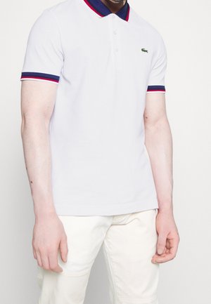 Homme portant un polo blanc avec des bordures bleu marine et rouges au col et aux manches, associé à un pantalon blanc cassé.