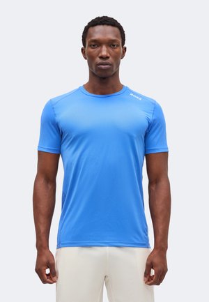 Férfi visel egy testhezálló, élénk kék, rövid ujjú sportpólót és világos színű nadrágot, előre nézve áll egy egyszínű háttér előtt.