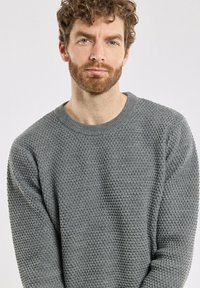 Un pull en tricot gris avec un motif texturé, un col rond et des manches longues, porté par une personne se tenant devant un fond uni.