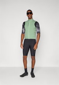 Radfahroutfit für Männer mit einem grünen, mit Reißverschluss versehenen Trikot und schwarzen sowie grauen gemusterten Ärmeln, kombiniert mit schwarzen Shorts und Radschuhen.