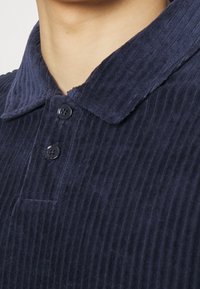 Chemise en velours côtelé bleu marine avec un col et deux boutons noirs visibles, portée par une personne montrée du cou au haut de la poitrine.