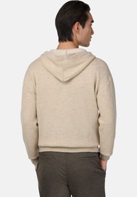 Hoodie beige à coupe décontractée, présentant une texture douce, une capuche avec cordon de serrage et des poignets côtelés. Associé à un pantalon gris foncé.