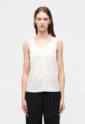 Γυναίκα με ίσια καστανά μαλλιά, που φοράει λευκό αμάνικο τοπ με V λαιμόκοψη και άνετο μαύρο παντελόνι, στέκεται μπροστά σε μια απλή φωτεινή επιφάνεια.