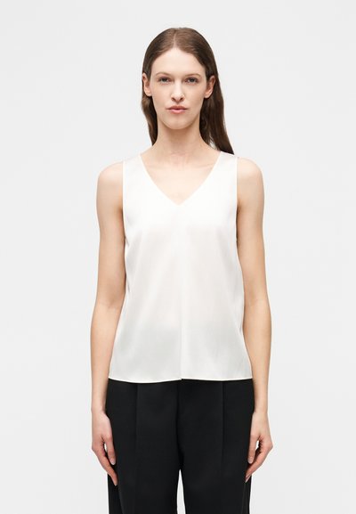 Femme aux cheveux bruns raides portant un haut blanc sans manches à encolure en V et un pantalon noir ample, debout devant un fond clair uni.