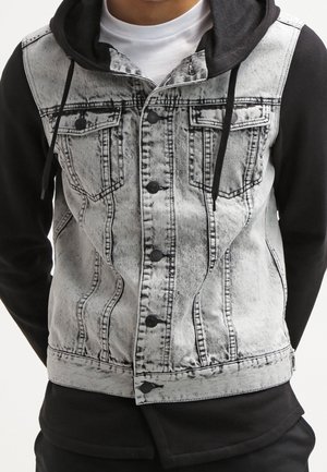 Homme portant un sweat à capuche noir sous une veste sans manches en denim délavé gris clair avec des boutons noirs et des poches poitrine.