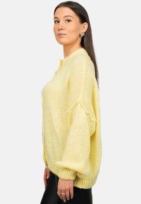 Cardigan tricoté jaune clair avec une coupe ample, des poignets côtelés et une fermeture à boutons. Présente un motif texturé en tricot ouvert.