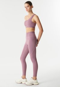 Paarse sportoutfit bestaande uit een sportbeha met één schouder en hoge taille leggings met naaddetails. Draagt een paar chunky witte sneakers.