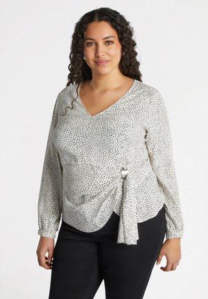 Blouse blanche à col en V avec des pois noirs, manches longues et un détail à nouer à la taille. Fabrication en tissu lisse et coupe décontractée.