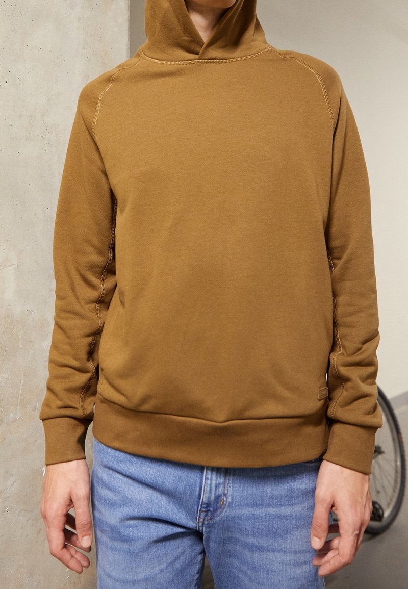 Sudadera marrón con capucha, con puños y dobladillo de canalé, hecha de una tela suave. Presenta costuras visibles y un corte holgado. Llevada sobre un vaquero azul claro.