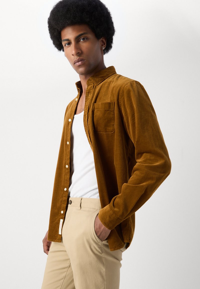 Braunes Cordhemd mit durchgehender Knopföffnung, einer einzelnen Brusttasche und einer weichen Textur, getragen über einem weißen Tanktop und beigen Hosen.