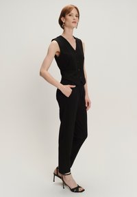 Schwarze ärmellose Weste mit V-Ausschnitt und Knopfverschluss, kombiniert mit maßgeschneiderten schwarzen Hosen. Bietet ein elegantes Design und eine schmale Silhouette.
