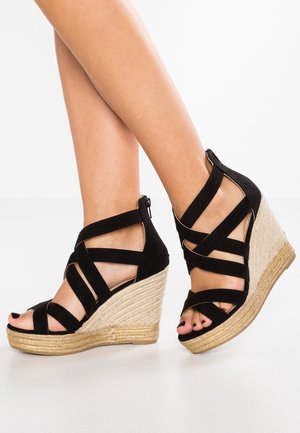 Ben iført sorte sandaler med remme, kilehæl i vævet espadrille-stil og åbne tæer på hvid baggrund.