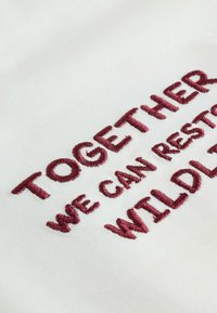 Tela blanca con texto bordado en color marrón que dice "JUNTOS PODEMOS RESTAURAR LA VIDA SILVESTRE." Detalle enfocado en la textura del bordado.