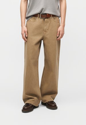 Persona che indossa pantaloni beige a taglio largo, cintura in pelle marrone, camicia grigia e scarpe marrone scuro, in piedi su uno sfondo chiaro e uniforme.