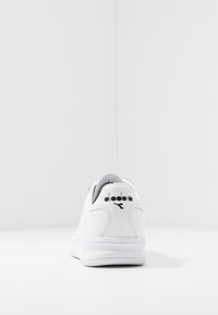 Sneaker bianco con tomaia in pelle liscia, forma arrotondata, suola bianca e marchio nero sul tallone. Design minimalista senza motivi visibili.