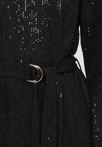 Svarte kjole med strukturt stoff med vertikale striper og spredte paljetter. Inkluderer et stoffbelte og en metallspenne.