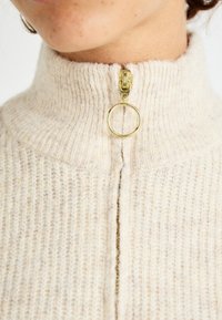 Cream knitted half-zip sweater with a high collar, featuring a gold zipper and circular pull, showcasing a textured pattern.