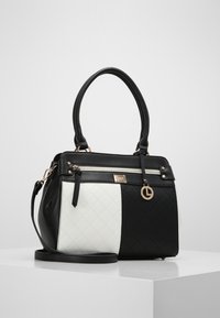 Mustek quiltingin musta-valkoinen käsilaukku, jossa on kaksinkertaiset kahvat, vetoketjullinen etutasku ja irrotettava olkahihna, asetettuna valkoiselle jalustalle.