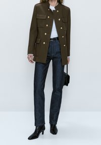 Veste militaire vert olive avec des boutons dorés, associée à un jean droit bleu foncé et des bottines noires. Design minimaliste.
