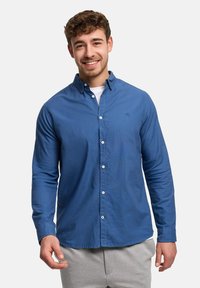 Leende man med lockigt hår, iklädd en blå skjorta med knappar över en vit underställströja och ljusgrå byxor, stående mot en enfärgad bakgrund.