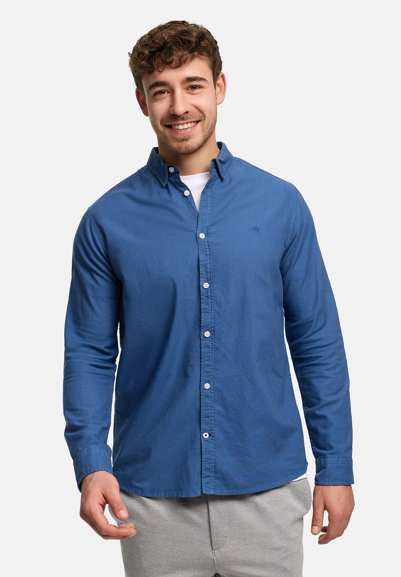 Leende man med lockigt hår, iklädd en blå skjorta med knappar över en vit underställströja och ljusgrå byxor, stående mot en enfärgad bakgrund.