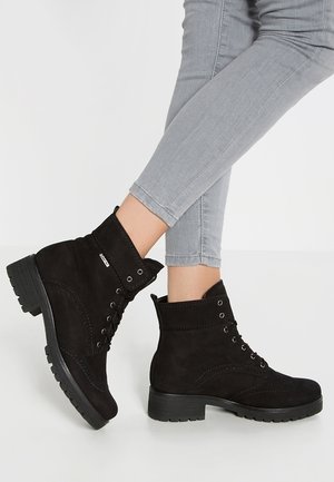 Botines negros de ante con cordones, usados con jeans ajustados gris claro, mostrando detalles laterales y de la suela con tacones gruesos y suelas con dibujo.
