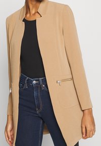 Beige sako z texturované tkaniny se střihem do rovna, s dvěma kapsami na zip, nošené přes černý top a tmavé džíny.