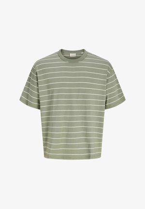 Garçon blond debout, portant une chemise à manches courtes rayée verte, un jean bleu ample et des baskets de couleur claire, souriant légèrement.