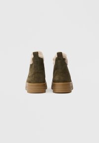 Olivově zelené semišové kotníkové boty s lehkým umělým kožešinovým podšívkou, elastickými bočními panely a texturovanými gumovými podrážkami. Zadní pohled ukazuje přímý siluetu.