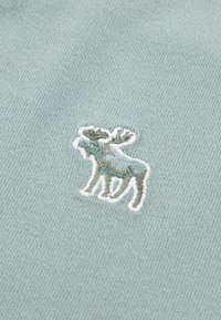 Un tissu bleu clair présente un orignal brodé en détail dans des nuances de vert et de marron, souligné en blanc, mettant en valeur la texture et le design.