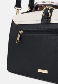 Sac à main noir texturé avec rabat supérieur blanc, fermeture éclair et ferrures dorées, et un détail en tissu imprimé léopard à l’intérieur de l’ouverture supérieure.