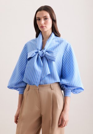 Femme portant un chemisier bleu clair à rayures blanches avec un grand nœud et des manches bouffantes, assorti à un pantalon taille haute beige.