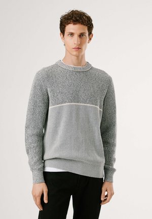 Grauer gestrickter Pullover mit einem strukturierten oberen Abschnitt, gerippten Bündchen und einem einfarbigen Streifen über der Brust. Über einem weißen Hemd getragen.