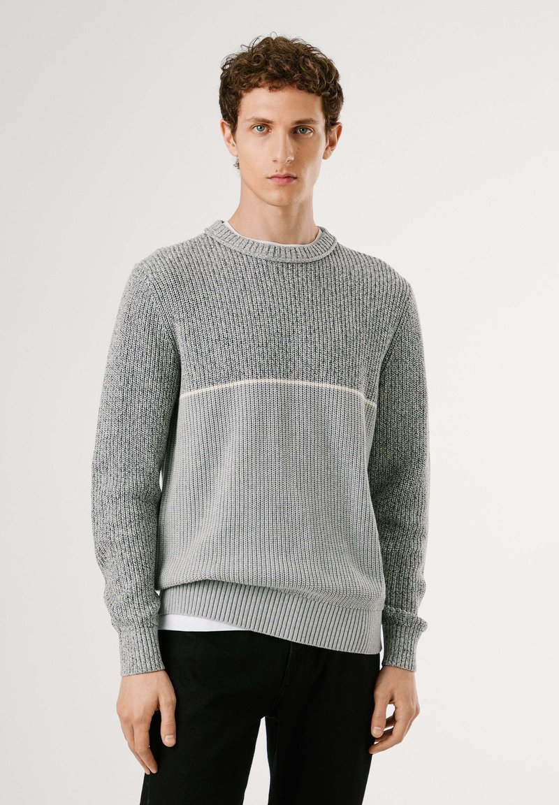 Grauer gestrickter Pullover mit einem strukturierten oberen Abschnitt, gerippten Bündchen und einem einfarbigen Streifen über der Brust. Über einem weißen Hemd getragen.