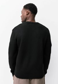 Zwarte gebreide trui met een gestructureerd oppervlak, geribbelde manchetten en zoom. Heeft een ronde halslijn en een losse pasvorm, gezien vanaf de achterkant.