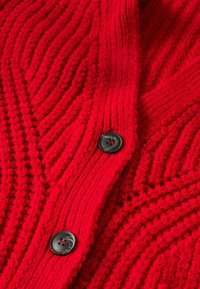 Punainen neulepusero, jossa on ribbipinta, erottuvat aaltoilevat kuviot ja mustat napit. Lähiotoksessa keskitytään kankaan ja nappien yksityiskohtiin.