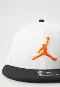 Hvid kasket med en sort skygge, der har et orange broderet Jumpman-logo. Materialet er glat stof med en struktureret design og flad skygge.